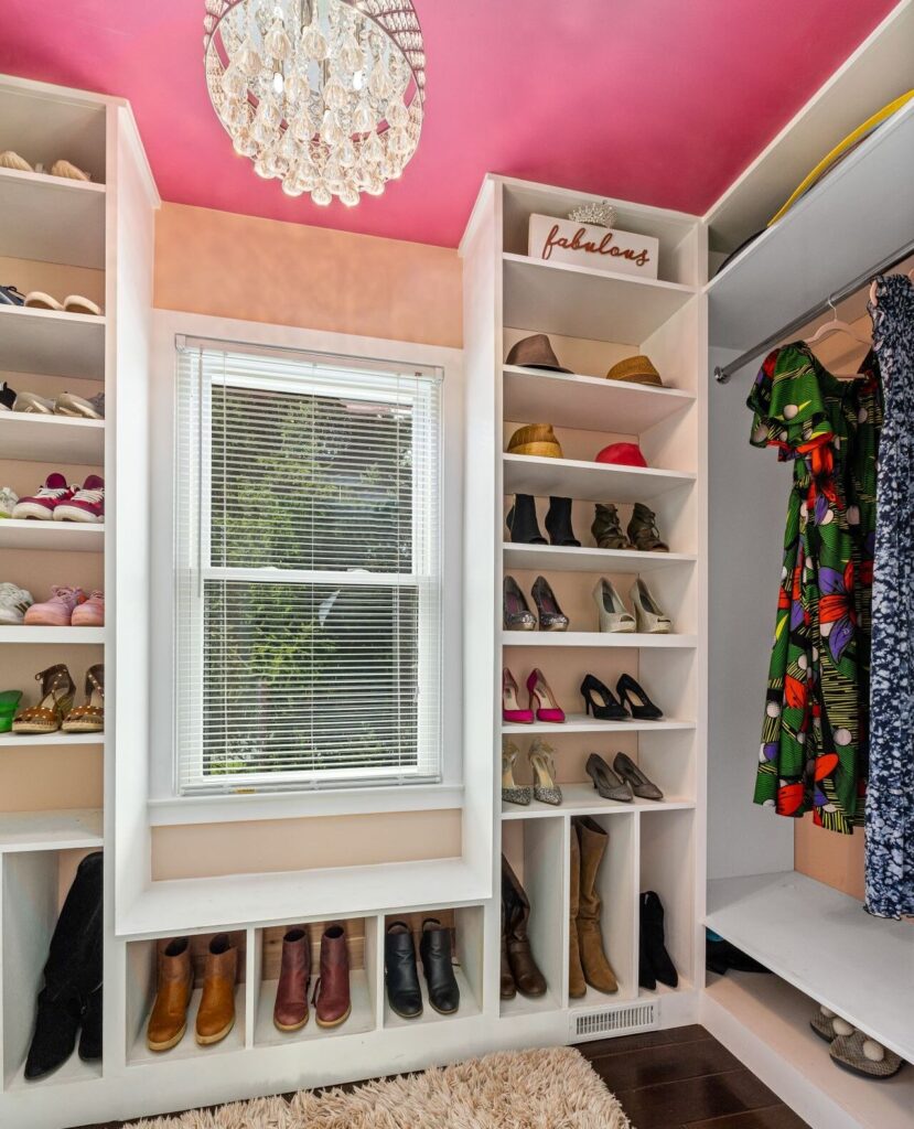 Organized Atlanta walk-in closet with a pink ceiling and chandelier, white built-in shelving displaying neatly arranged shoes, hats, boots, and hanging dresses beside a window